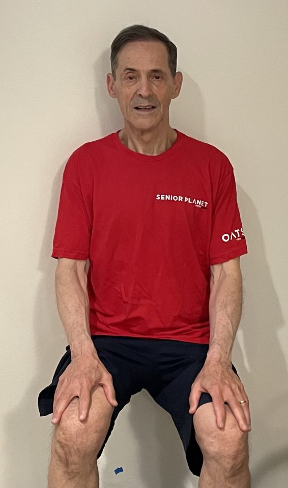 man in red Senior Planet shirt doing a wall-sit exerciese