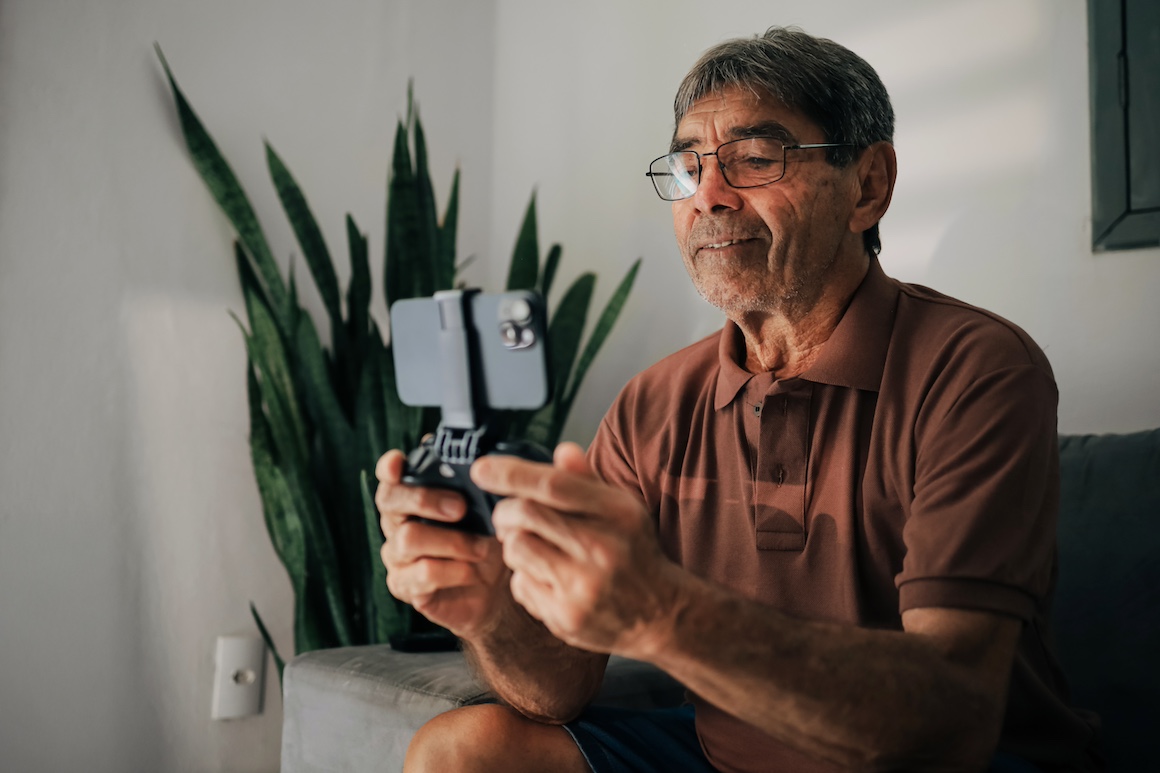 Senior man focused on playing mobile game with smartphone controller while sitting on couch at home